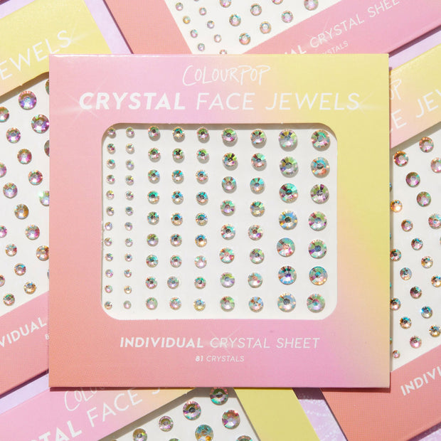 ColourPop Crystal Face Jewels Individual crystal sheet showcasing 81 iridescent adhesive gems in multiple sizes arranged in neat rows, set in a pink-to-yellow gradient package on a flat lay of overlapping boxes.