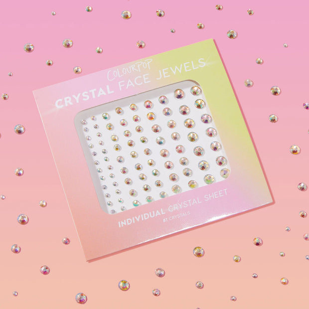ColourPop Crystal Face Jewels Individual sheet displaying 81 adhesive iridescent rhinestones arranged by size, shimmering in a die-cut window, set against a pink gradient background with scattered crystals for a sparkly makeup look.