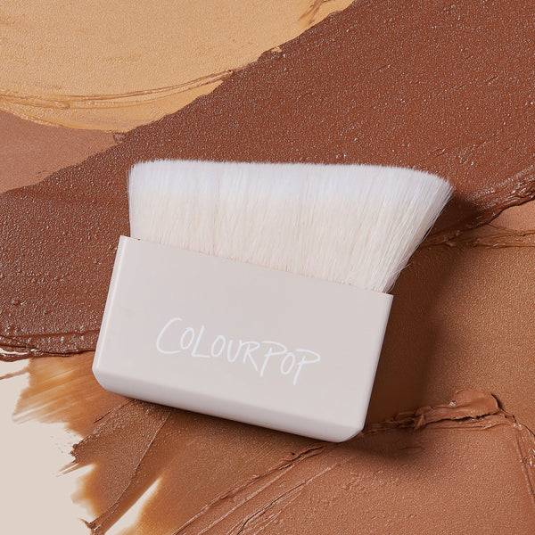 ColourPop Makeup Brush Angled Sculpting Brush resting on swatches of tan to deep brown cream contour; dense white angled bristles extend from a beige block handle stamped ColourPop, ideal for precise cheekbone sculpting.
