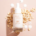 Fourth Ray Beauty Oat Face Milk dropper bottle standing amid oat flakes, with milky serum drops on a nude background; hydrating ColourPop Face Milk highlights a gentle oat formula and creamy texture for soothing skincare.