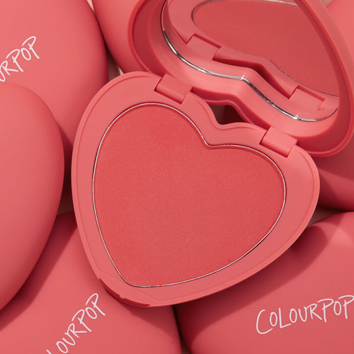 4ever Yours ColourPop Pressed Powder Blush compact opens to reveal a heart-shaped coral pan and mirror, set atop several matte pink cases with ColourPop logos, highlighting the romantic, travel-friendly pressed powder blush.
