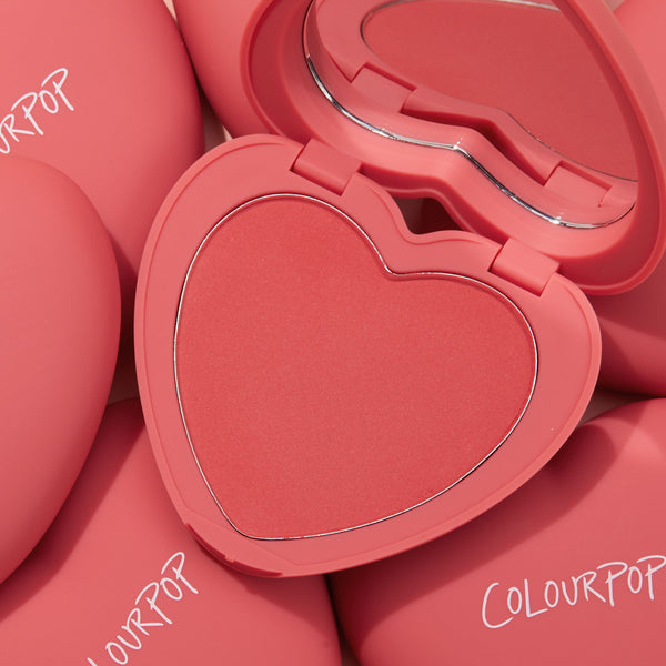 4ever Yours ColourPop Pressed Powder Blush compact opens to reveal a heart-shaped coral pan and mirror, set atop several matte pink cases with ColourPop logos, highlighting the romantic, travel-friendly pressed powder blush.