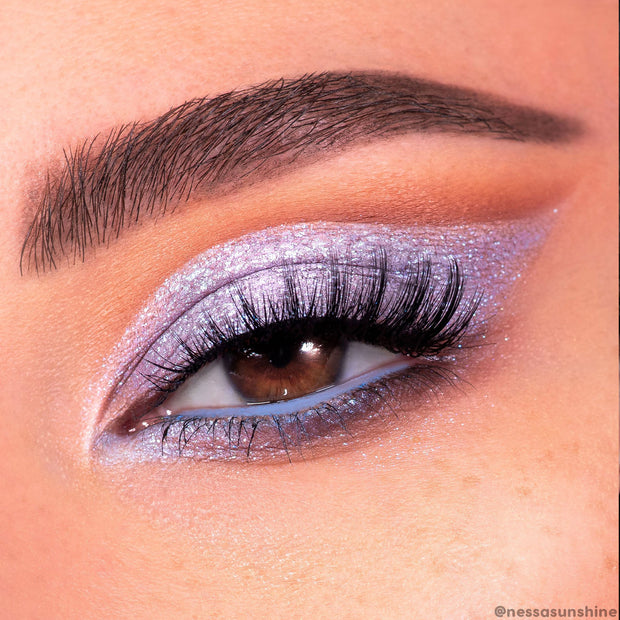 Moon Daisy Super Shock Shadow by ColourPop shimmering lilac on eyelid, blended into a wing with icy lavender sparkle and inner-corner highlight, paired with dramatic lashes, soft peach crease, and periwinkle liner on the lower lash line.