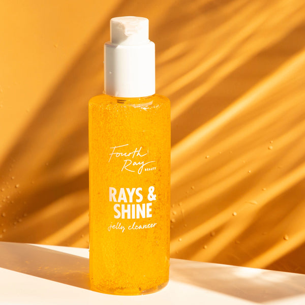 Rays and Shine Cleanser jelly cleanser bottle from Fourth Ray Beauty by ColourPop stands upright, amber gel with bubbles glinting in sunlight, on a white ledge against a warm orange backdrop with soft shadows and water droplets.