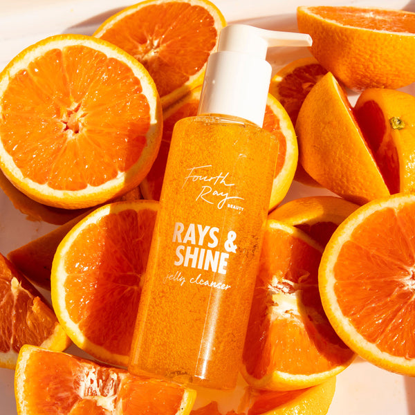 Rays and Shine Cleanser jelly cleanser by ColourPop, a Fourth Ray Beauty pump bottle resting among juicy orange halves on a sunlit white surface, showcasing bright citrus-orange gel and sleek, glossy skincare packaging.