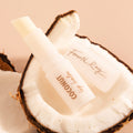 Coconut Lip Balm by Fourth Ray Beauty for ColourPop, tube uncapped to reveal creamy stick, nestled inside a halved coconut on a warm beige backdrop.