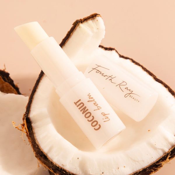 Coconut Lip Balm by Fourth Ray Beauty for ColourPop, tube uncapped to reveal creamy stick, nestled inside a halved coconut on a warm beige backdrop.