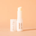 ColourPop Fourth Ray Beauty Coconut lip balm standing uncapped, creamy balm bullet exposed in a glossy white tube labeled COCONUT lip balm, cap beside it, on a warm peach background with soft shadow.