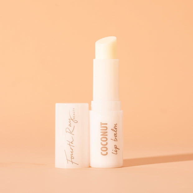 ColourPop Fourth Ray Beauty Coconut lip balm standing uncapped, creamy balm bullet exposed in a glossy white tube labeled COCONUT lip balm, cap beside it, on a warm peach background with soft shadow.