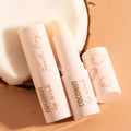 Coconut Lip Scrub stick by Fourth Ray Beauty, opened to show the balm, resting beside its cap with coconut icons on a halved coconut against a warm beige backdrop; gentle exfoliating, hydrating treatment, ColourPop Lip Scrub.