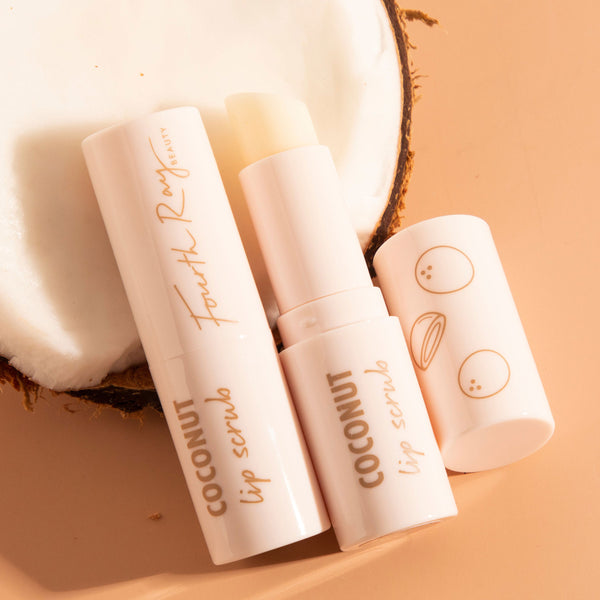 Coconut Lip Scrub stick by Fourth Ray Beauty, opened to show the balm, resting beside its cap with coconut icons on a halved coconut against a warm beige backdrop; gentle exfoliating, hydrating treatment, ColourPop Lip Scrub.