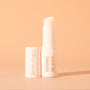 Coconut ColourPop Lip Scrub stick stands upright with the cap off, revealing an angled balm in a sleek ivory Fourth Ray Beauty tube with gold “COCONUT lip scrub” text on a soft peach background.