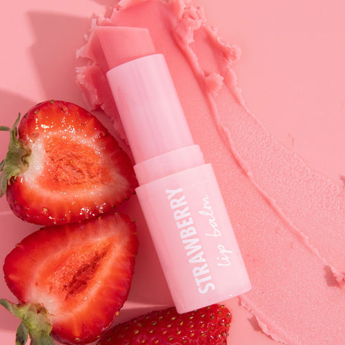 Strawberry lip balm in a pastel pink twist-up tube labeled STRAWBERRY lies open with the slanted balm exposed, beside a creamy swipe, framed by fresh strawberry halves on a pink backdrop; ColourPop Lip Balm, fruity and moisturizing.