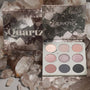 ColourPop Of Quartz shadow palette showcases nine cool-toned pressed powder shades - a mix of shimmers and mattes - in an open 3x3 compact beside its crystal-print box, arranged among smoky quartz rocks.
