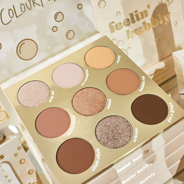 ColourPop Feelin’ Bubbly shadow palette showcases nine neutral champagne, bronze, and brown shades in matte and shimmer finishes, arranged in a gold 9-pan layout, open on a stack of palettes with bubbly motifs and foil branding.