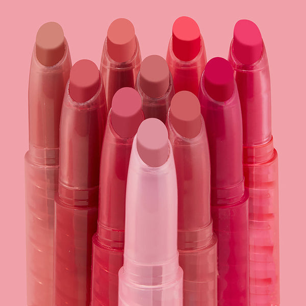 Just A Kiss ColourPop Lippie Stix set standing upright in a tight cluster, showcasing creamy bullet tips in nude, pink, coral, and berry tones, in translucent pink twist-up tubes against a soft blush background.