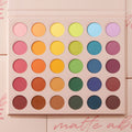 Matte About Hue ColourPop Shadow Palette displays 30 matte rainbow shadows - pink, orange, yellow, green, blue, purple, and neutrals - arranged in five rows of round pans, opened in a blush-pink case with printed shade names on a soft pink background.