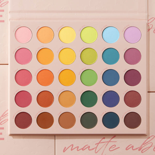 Matte About Hue ColourPop Shadow Palette displays 30 matte rainbow shadows - pink, orange, yellow, green, blue, purple, and neutrals - arranged in five rows of round pans, opened in a blush-pink case with printed shade names on a soft pink background.