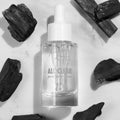 Glass dropper bottle of Fourth Ray Beauty All Clear Purifying Serum standing upright, showcasing clear formula and label, surrounded by charcoal pieces on a white marble surface; .85 fl oz/25 mL ColourPop Face Serum for purifying skin.