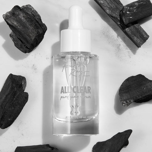 Glass dropper bottle of Fourth Ray Beauty All Clear Purifying Serum standing upright, showcasing clear formula and label, surrounded by charcoal pieces on a white marble surface; .85 fl oz/25 mL ColourPop Face Serum for purifying skin.
