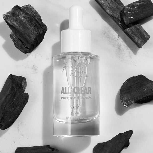Glass dropper bottle of Fourth Ray Beauty All Clear Purifying Serum standing upright, showcasing clear formula and label, surrounded by charcoal pieces on a white marble surface; .85 fl oz/25 mL ColourPop Face Serum for purifying skin.
