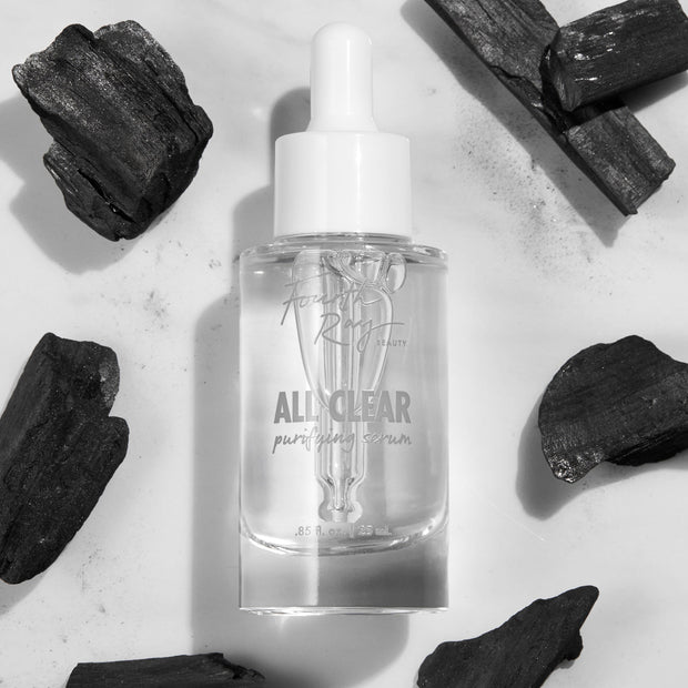 Glass dropper bottle of Fourth Ray Beauty All Clear Purifying Serum standing upright, showcasing clear formula and label, surrounded by charcoal pieces on a white marble surface; .85 fl oz/25 mL ColourPop Face Serum for purifying skin.