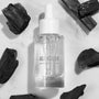 Glass dropper bottle of Fourth Ray Beauty All Clear Purifying Serum standing upright, showcasing clear formula and label, surrounded by charcoal pieces on a white marble surface; .85 fl oz/25 mL ColourPop Face Serum for purifying skin.