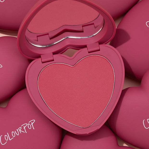 Heart-shaped compact in shade Babycakes opens to reveal a rosy pink ColourPop Pressed Powder Blush with a heart-shaped mirror, surrounded by matching magenta heart cases with ColourPop branding.