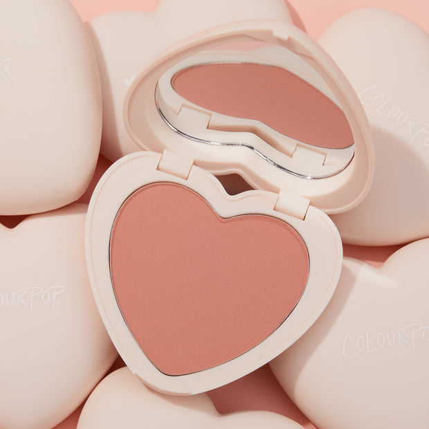 ColourPop Kiss 'N Tell pressed powder blush in a heart-shaped compact, opened to reveal a matte peachy‑pink pan and mirror, sitting among cream heart compacts on a soft pink backdrop.