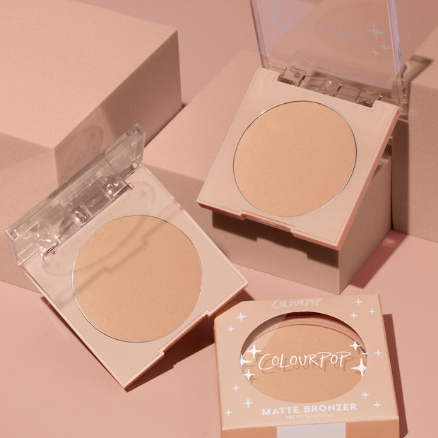 Avila Beach ColourPop Pressed Powder Bronzer compact opens to reveal a warm matte tan pan, with a second open compact and boxed matte bronzer nearby on soft pink blocks, showcasing clear flip-top lids and the 6.0 g pressed powder.