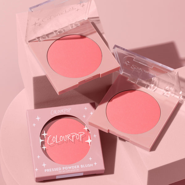 ColourPop Pressed Powder Blush Big Reveal compact displays a warm coral‑pink pan as two clear‑lid cases sit open and one boxed unit rests on a blush‑toned podium, star accents on the lids catching light for a soft, glam display.