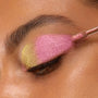 Doe-foot applicator sweeping ColourPop Chrome Liquid Shadow in Bliss across a closed eyelid, showing a metallic duochrome shift from lime-gold at the inner corner to vivid pink over the lid in a close-up beauty shot.