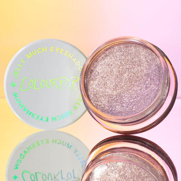ColourPop Jelly Much Shadow Boo-Kay shown open, a sparkling lavender-lilac gel eyeshadow in a clear round pot, white lid with neon green logo beside it, reflected on a mirror with a pink-to-yellow gradient background.