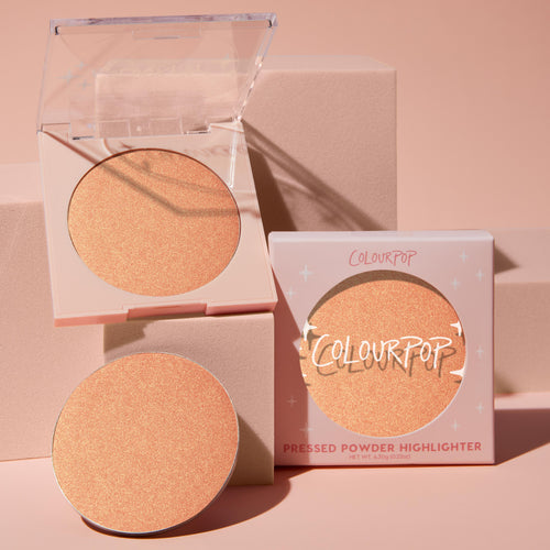 Boombox pressed powder highlighter catching light in an open compact, single refill pan, and boxed packaging, its peachy‑gold shimmer glowing; ColourPop Pressed Powder Highlighter styled on soft pink blocks in a studio setup.