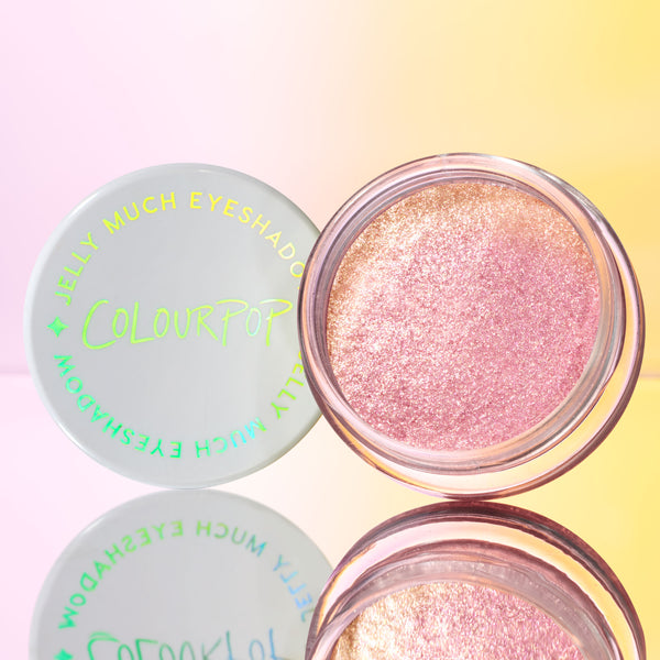 Open jar of ColourPop Jelly Much Shadow, shade Close To You, displays a shimmery pink-gold gel eyeshadow in a clear pot, with a white lid and neon green logo beside it, reflecting on a mirror against a pink-to-yellow backdrop.