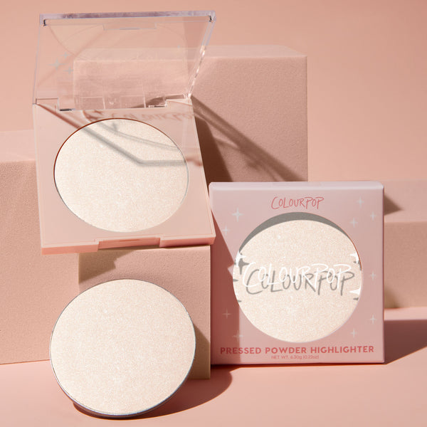 Cold One highlighter compact opens to reveal an icy pearl 0.23 oz pan, with a refill and star-print box beside it; ColourPop Pressed Powder Highlighter in a pale pink case styled on blush-pink blocks for a luminous glow.