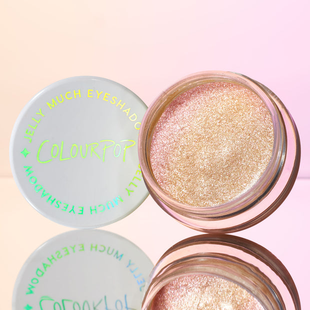 Open pot of ColourPop Jelly Much Shadow, Field of Fairies, revealing champagne‑pink, metallic glitter jelly eyeshadow; white lid with neon green ColourPop logo beside it on a mirrored surface against a soft pink gradient background.