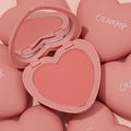Heart-shaped Let’s Dance ColourPop Pressed Powder Blush compact opens to reveal a coral pink powder pan and built-in mirror, resting among matte pink ColourPop heart cases on a soft blush backdrop.