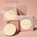 Minted pressed powder highlighter pan glows in an open clear-lid compact beside a refill and boxed packaging, showcasing the pale champagne-gold shade of the ColourPop Pressed Powder Highlighter on soft pink blocks under warm studio light.