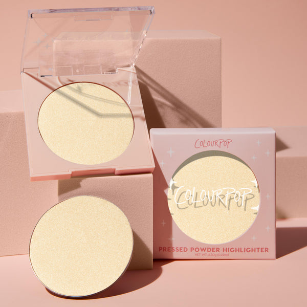 Minted pressed powder highlighter pan glows in an open clear-lid compact beside a refill and boxed packaging, showcasing the pale champagne-gold shade of the ColourPop Pressed Powder Highlighter on soft pink blocks under warm studio light.