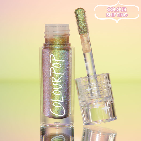 See U Later ColourPop Chrome Liquid Shadow stands open, its doe-foot applicator showing a green-gold to lavender color-shifting shimmer, set on a reflective surface with a pastel gradient background in a clear tube with white ColourPop logo.