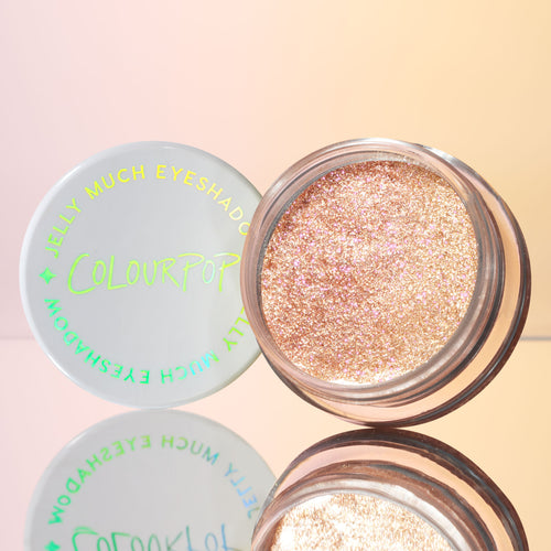 Open jar of ColourPop Jelly Much Shadow in Mystical displays a high-shine champagne‑pink glitter gel eyeshadow, set on a mirrored surface with the white lid and neon logo beside it against a soft peach pastel background.
