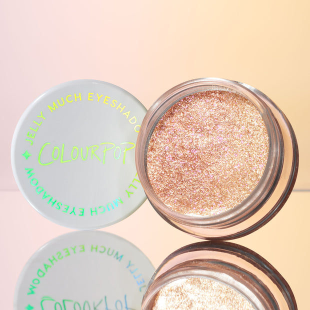 Open jar of ColourPop Jelly Much Shadow in Mystical displays a high-shine champagne‑pink glitter gel eyeshadow, set on a mirrored surface with the white lid and neon logo beside it against a soft peach pastel background.