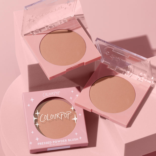 ColourPop Pressed Powder Blush in Out & About displayed in pink compacts with clear flip lids, warm peach‑nude matte pans, and one boxed unit, arranged on soft pink blocks in a clean product flat lay.