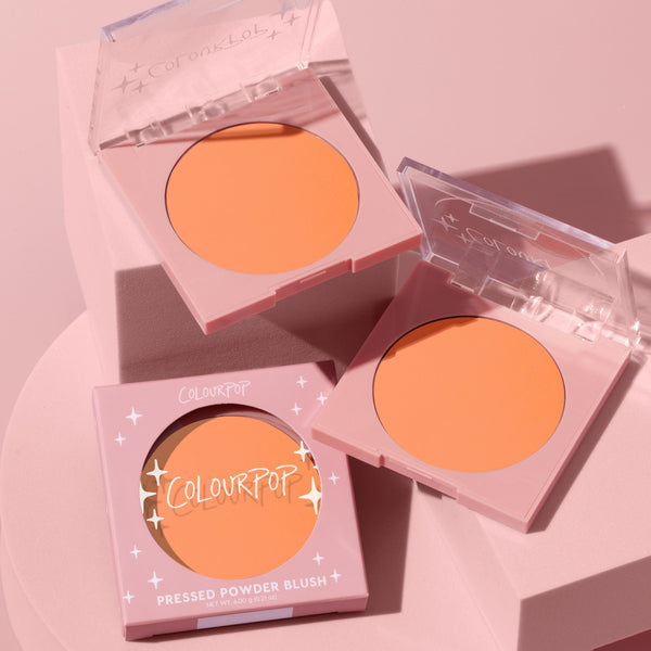 Roxbury Dr compact displays a matte peachy-coral pan, with two open cases and one boxed, arranged on pink geometric blocks in soft studio light; ColourPop Pressed Powder Blush in pastel pink packaging with clear lids and star accents, 4.00 g/0.14 oz.