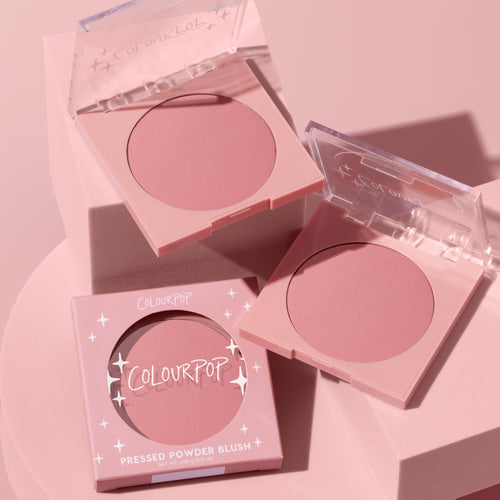 ColourPop Pressed Powder Blush in Sassy Pants, a matte dusty-rose hue, displayed in two open pastel-pink square compacts with clear lids beside the boxed unit with star accents, styled on a monochromatic blush-pink pedestal.