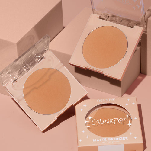 ColourPop Pressed Powder Bronzer in Silver Strand Beach, a warm matte tan shade, sits open in a peach compact with clear lid, displayed with a second open pan and boxed unit on soft pink blocks in a clean studio setup.