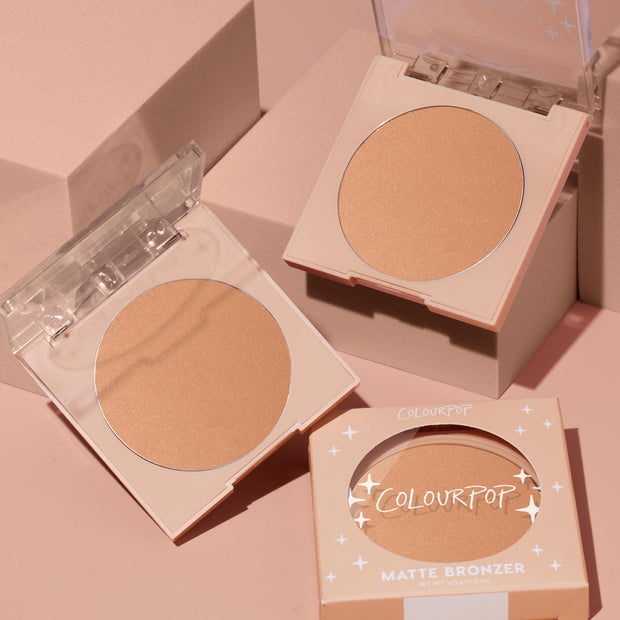ColourPop Summerland Beach pressed powder bronzer compact sits open to reveal a warm matte tan pan, alongside a second open compact and a boxed unit on blush‑pink blocks; ColourPop Pressed Powder Bronzer for a natural sun‑kissed finish.