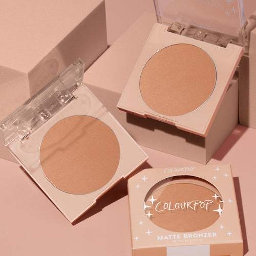 ColourPop Pressed Powder Bronzer Talk to the Palm compacts lying open to reveal a warm matte tan pan, with a boxed compact in starry peach packaging and clear lids, styled on blush-pink geometric blocks.