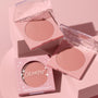 To the 10 ColourPop Pressed Powder Blush in pastel pink compacts, opened to show a soft rosy nude shade, styled with its boxed packaging on a pink pedestal under soft studio lighting, 6.0 g (0.21 oz) pan.
